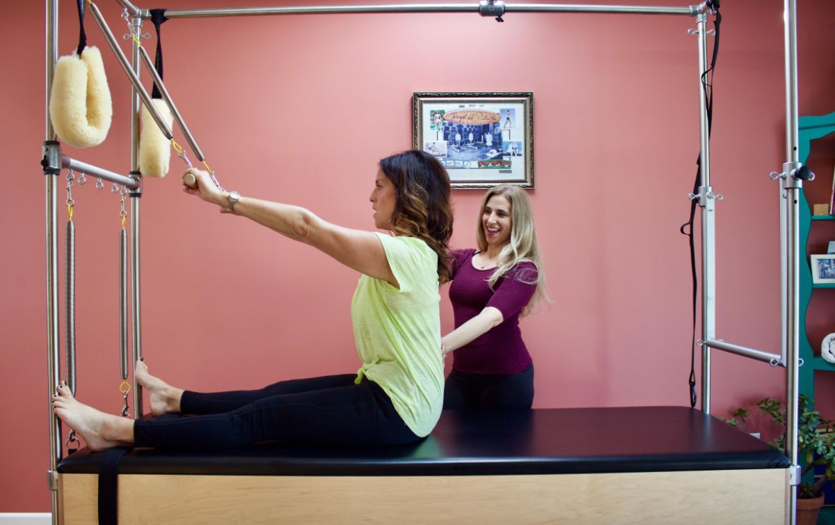 Woman exercising on Pilates reformer as trainer assists in a pink studio