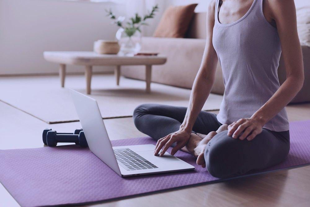 Person meditating on a yoga mat beside an open laptop in a bright room