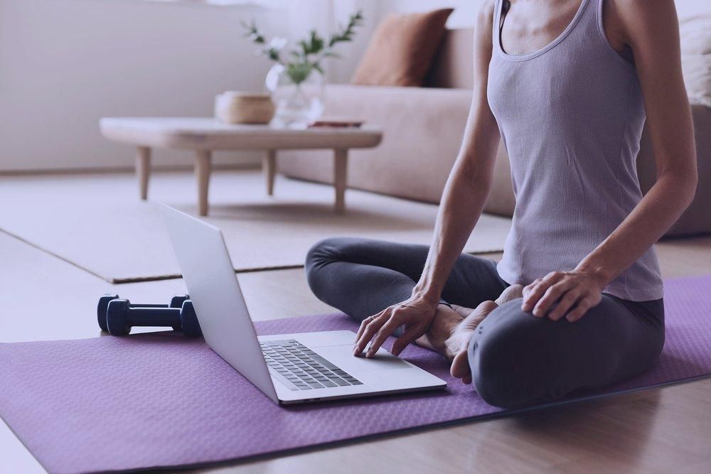 Woman in workout clothes, seated with laptop on yoga mat, in a living room.