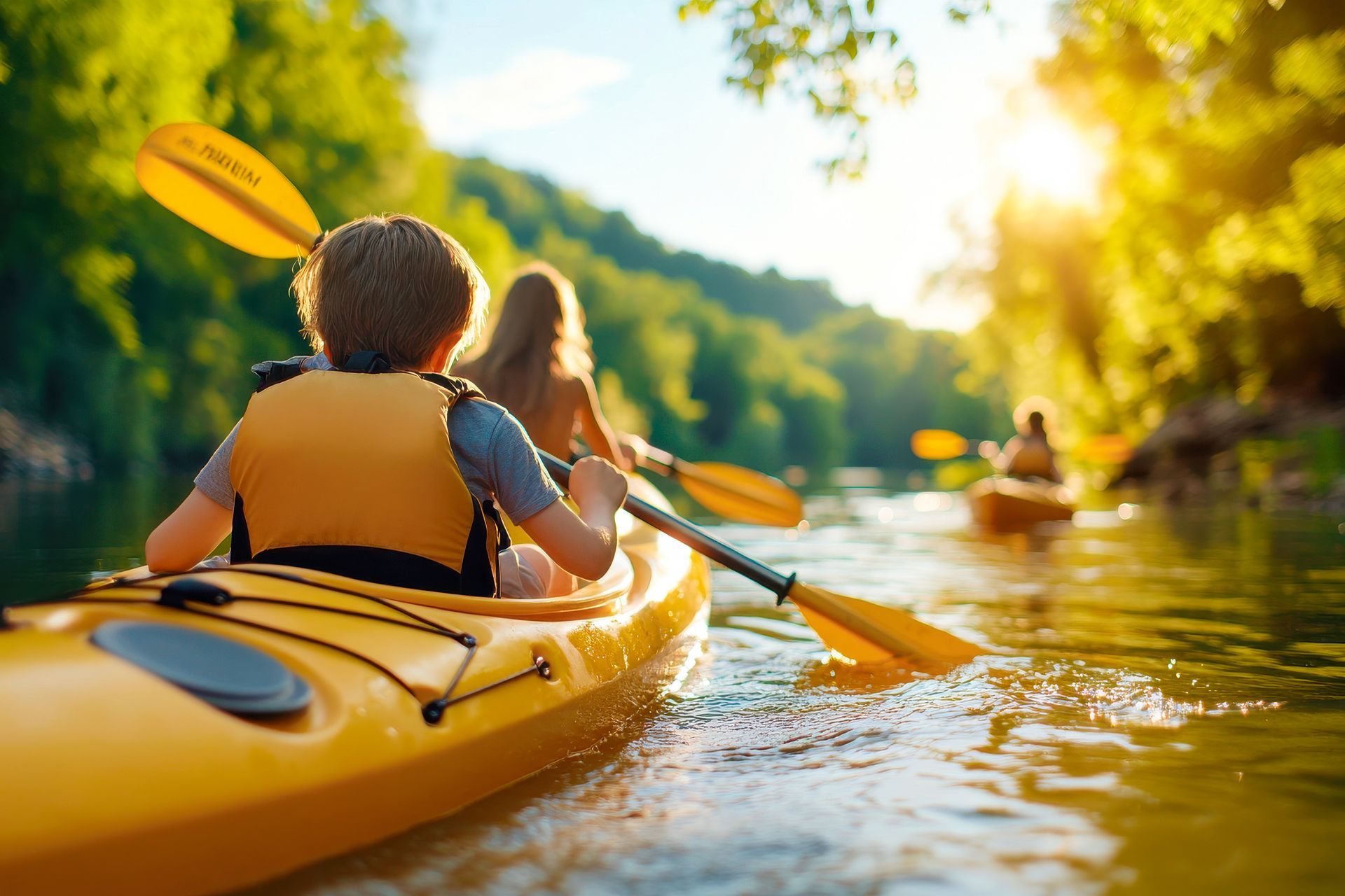 Two kayakers paddling a yellow kayak on a sunny river surrounded by green trees