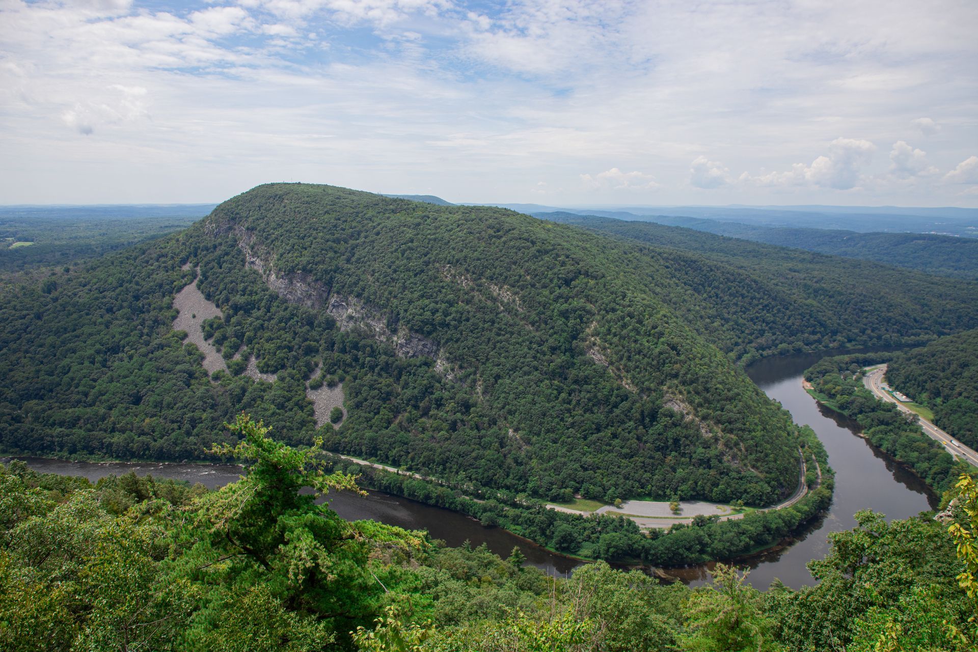 A winding river cuts through lush green forested hills under a partly cloudy sky.