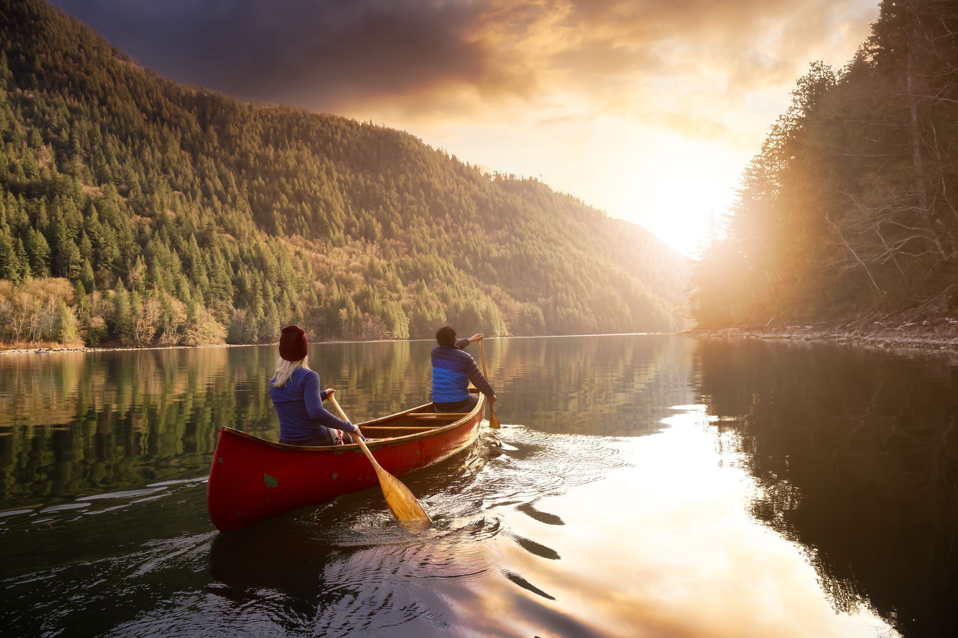 Two people canoe on a calm lake at sunrise, with forested mountains and golden light отражing on the water.