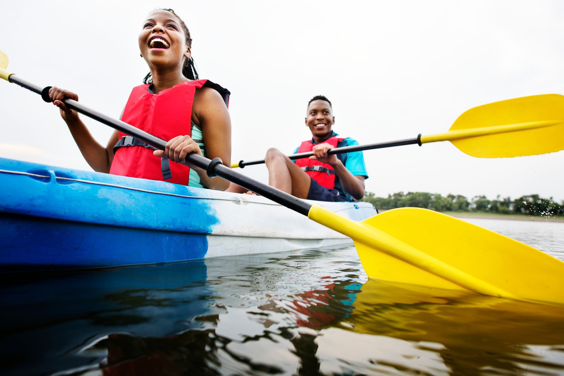 Two kayakers in red life vests paddling colorful boats on calm water, smiling outdoors