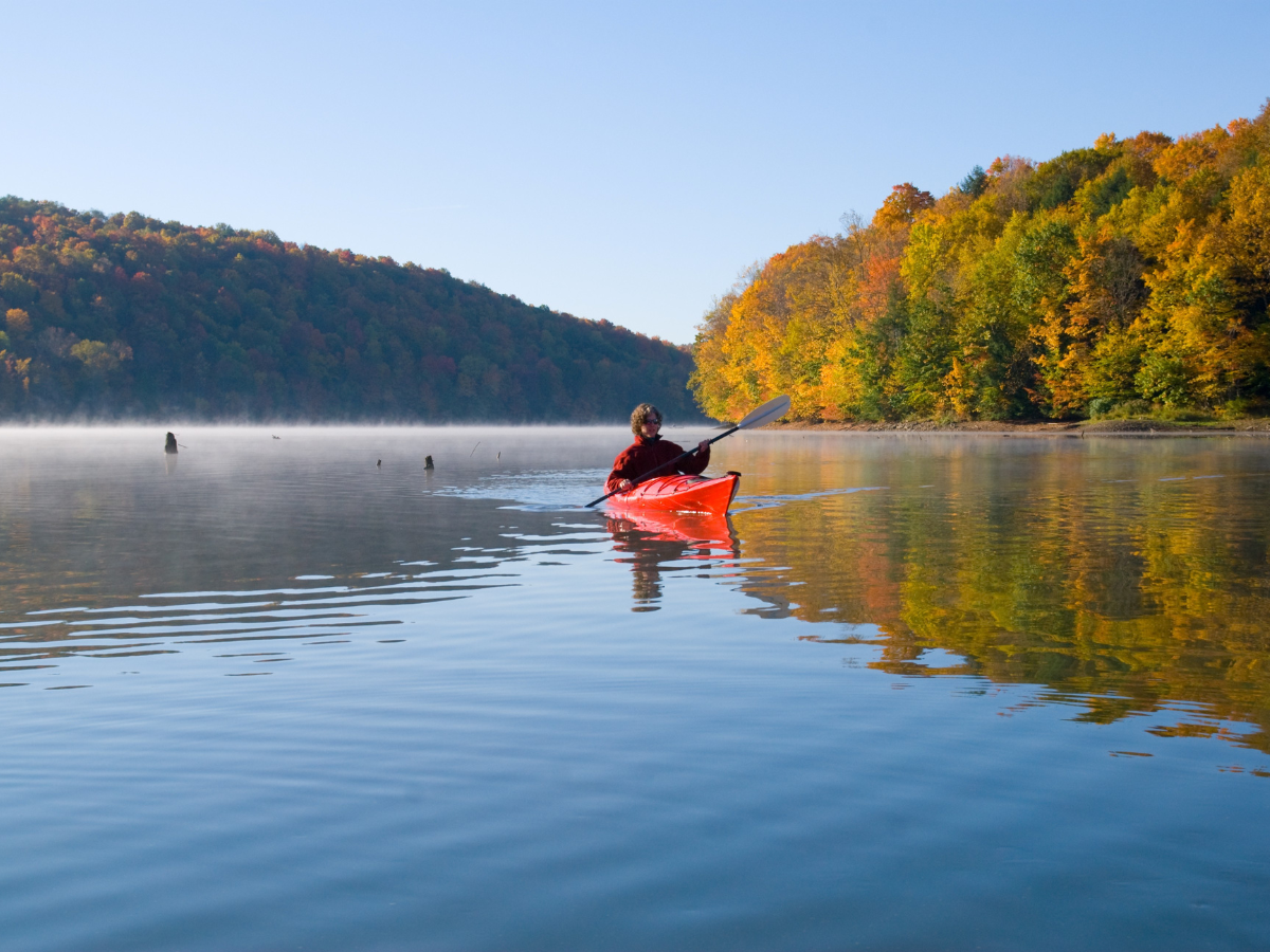 Kayaker paddling on calm lake; autumn foliage in background.
