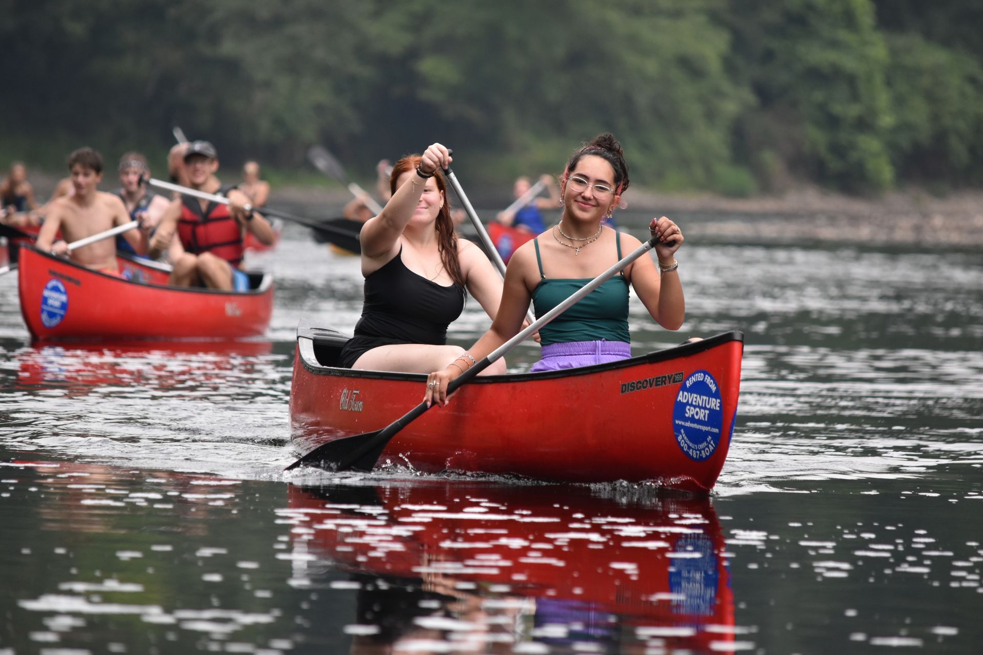 Two people paddling red canoes on a calm river with other canoers in the background.