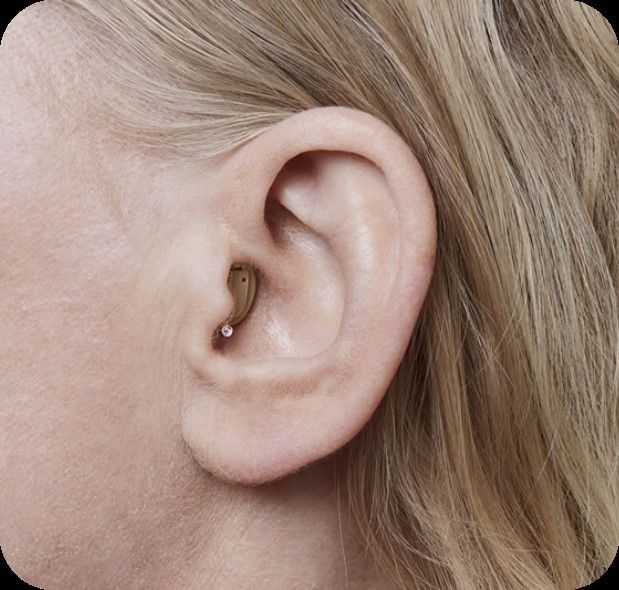 Primer plano de la oreja de una persona con un pequeño audífono beige dentro. La persona tiene cabello rubio.