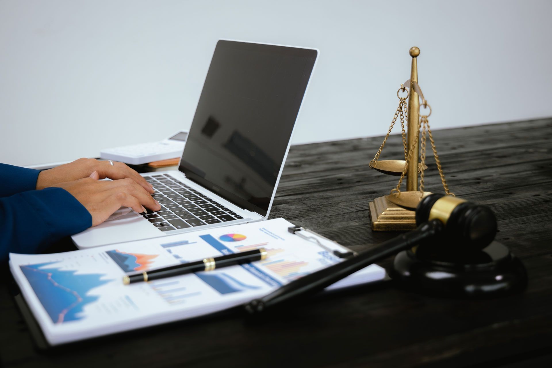 Hands typing on laptop beside papers, pen, headphones, and a brass balance scale on a desk