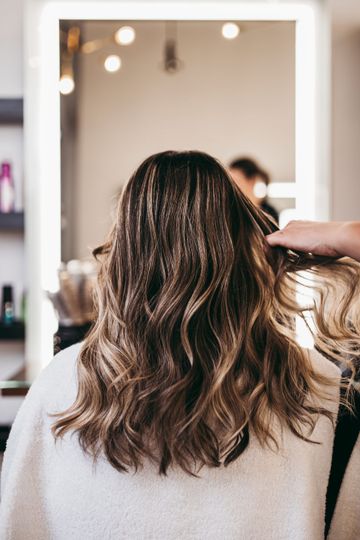 Person with wavy brown hair sits in a salon chair, facing a mirror while styling is done.