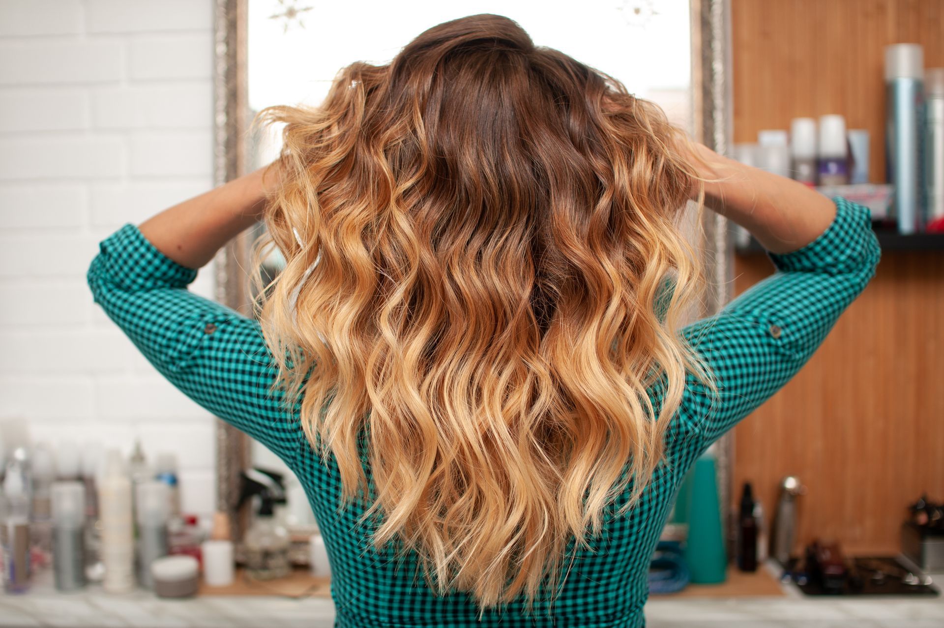 Back view of person with long wavy ombré hair, hands lifting hair in a bathroom mirror area