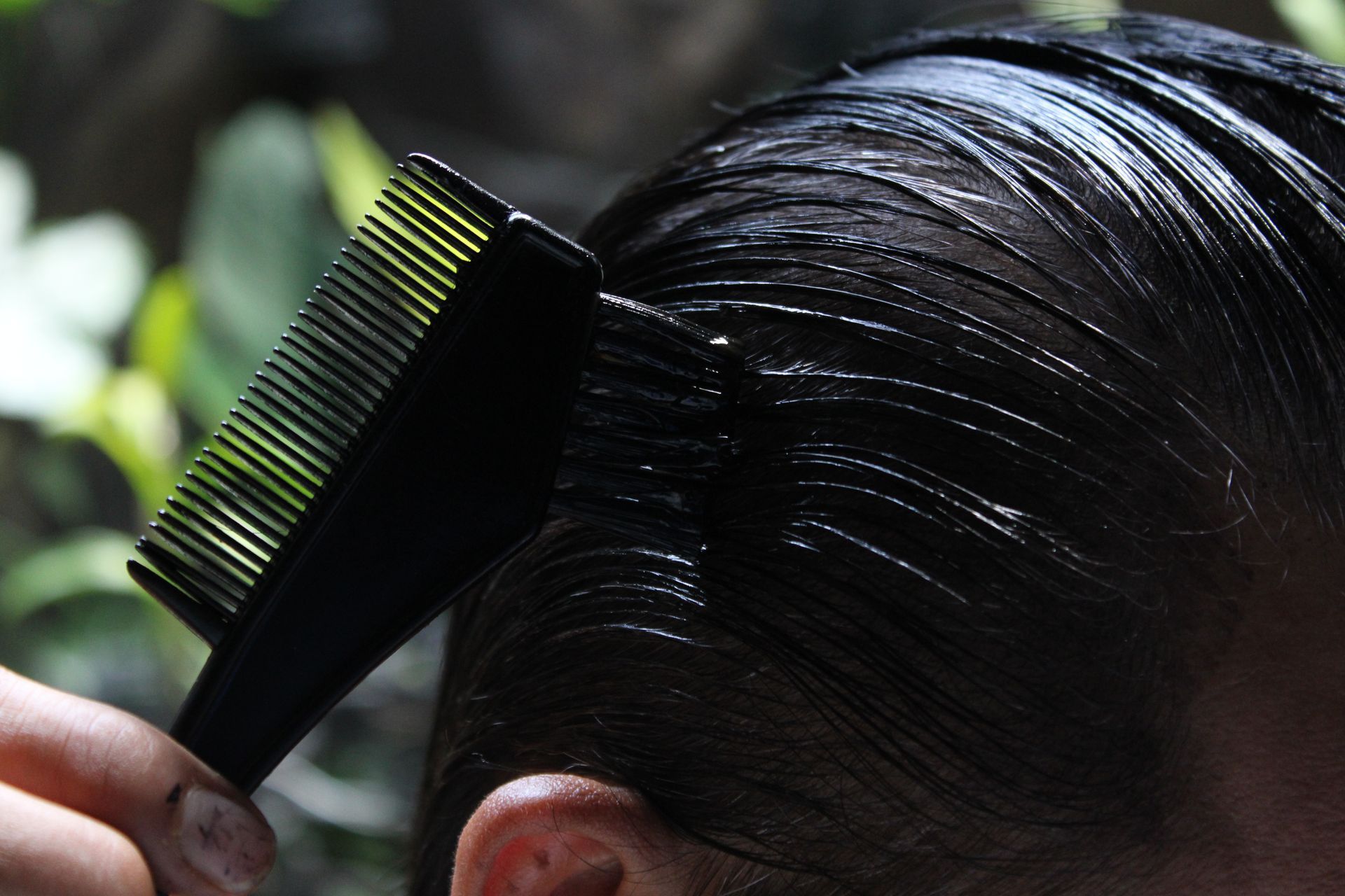 A hand combs through sleek black hair with a fine-tooth comb outdoors.