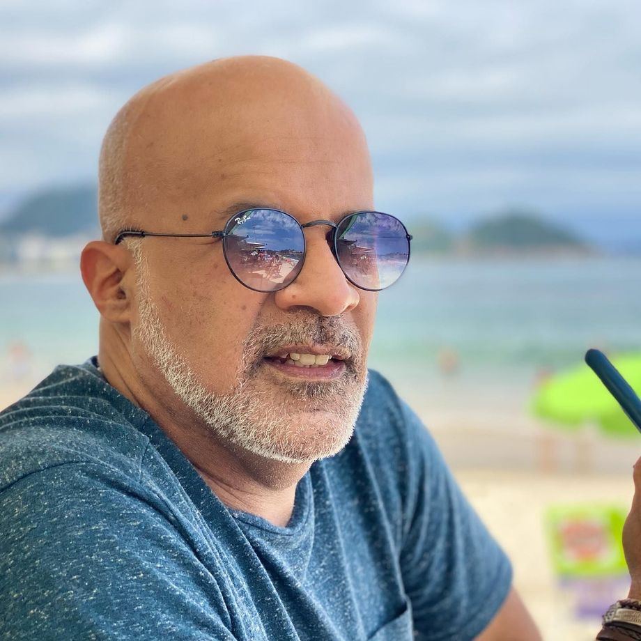 Um homem careca com barba usando óculos escuros e uma camisa azul