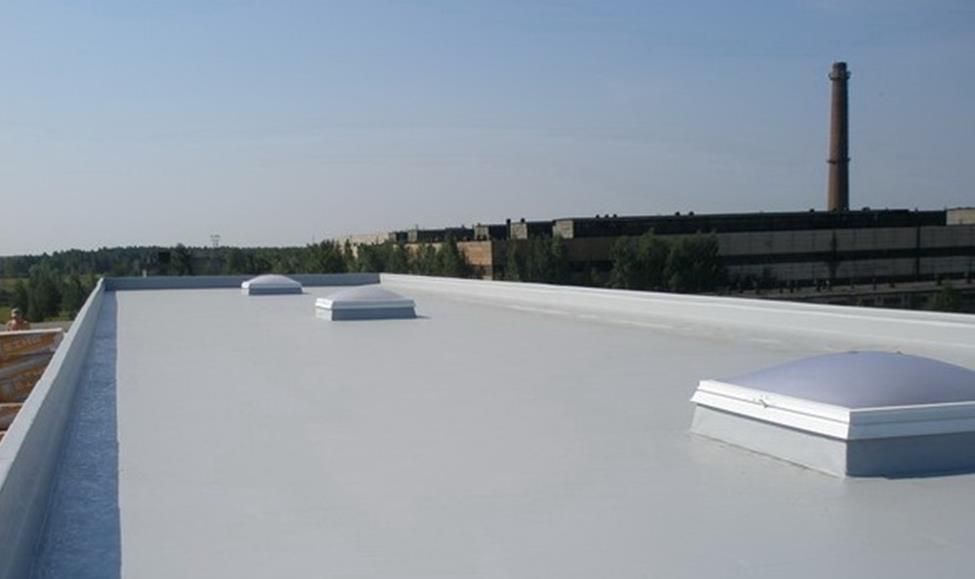 A view across a flat, light-gray roof with three skylights, showing an industrial building and chimney in the background.