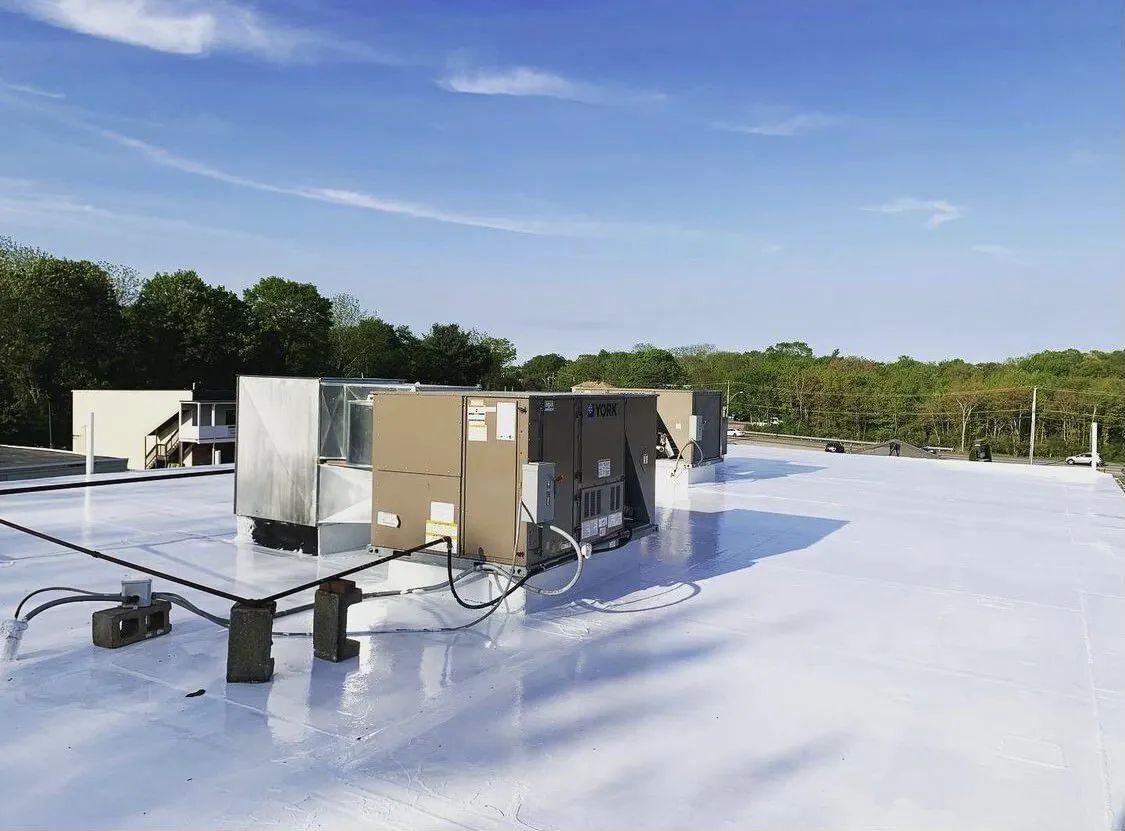 White-coated flat roof with several HVAC units. Bright blue sky with trees in the background.