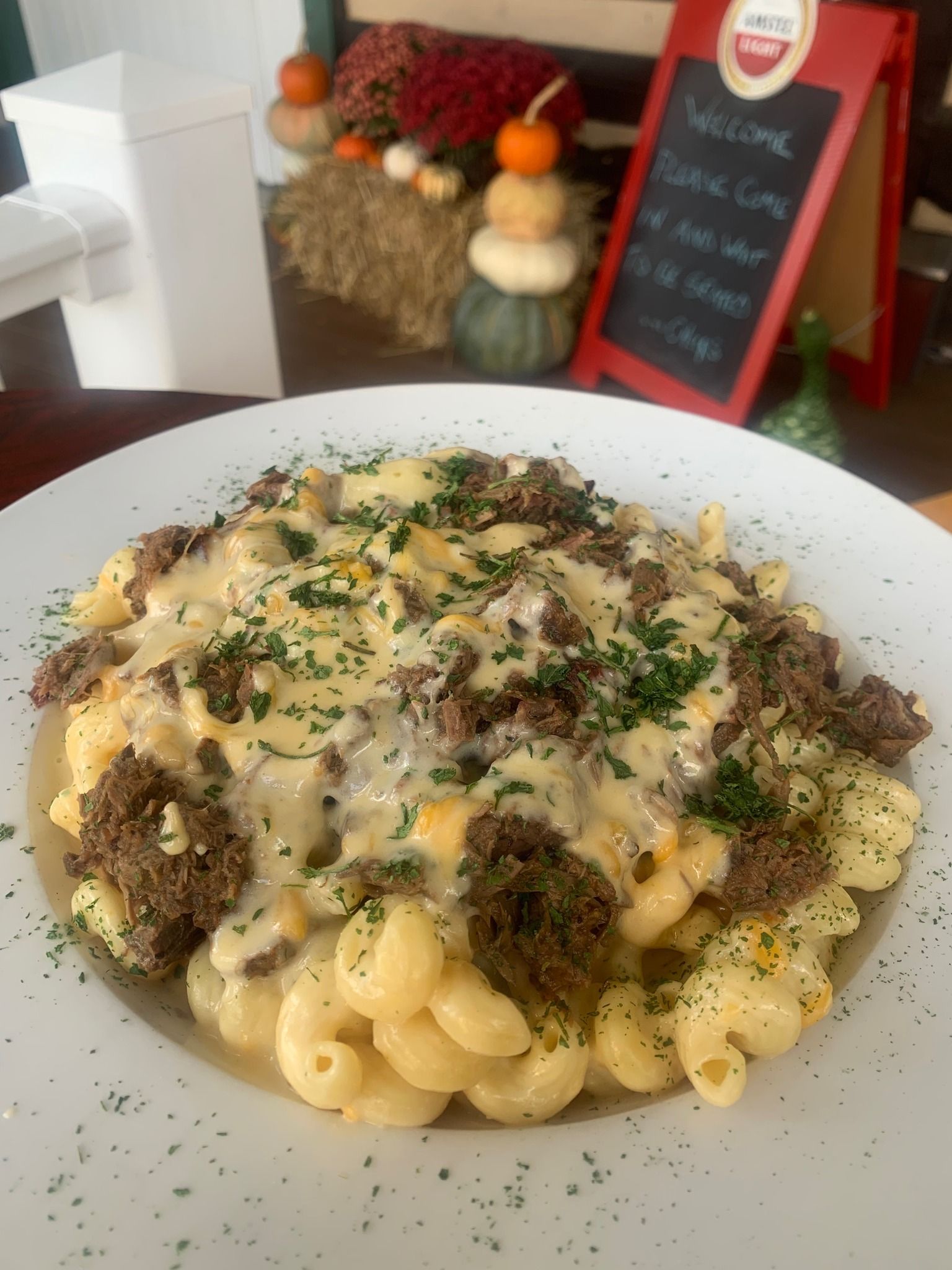 Creamy pasta topped with herb-crusted meat on a white plate, with a small chalkboard menu in the background