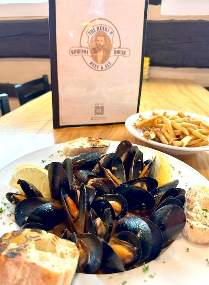 Mussels in a white bowl with fries and bread on a restaurant table, with a menu in the background