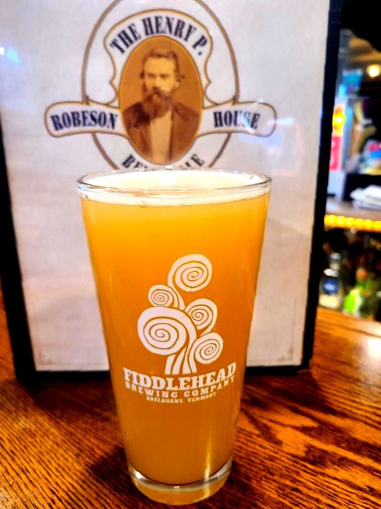 Pint of hazy orange beer on a wooden table, with a brewery logo in the background