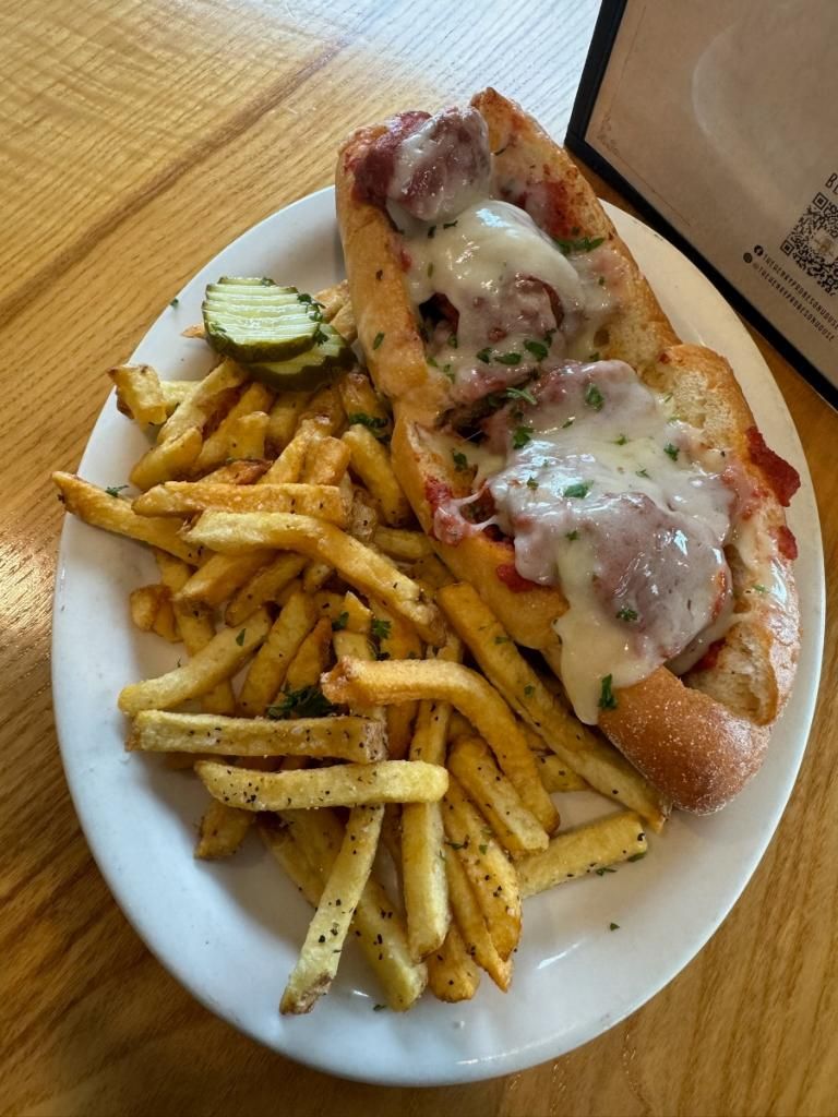 Loaded hot dog with melted cheese, fries, and a pickle on a white plate at a wooden table