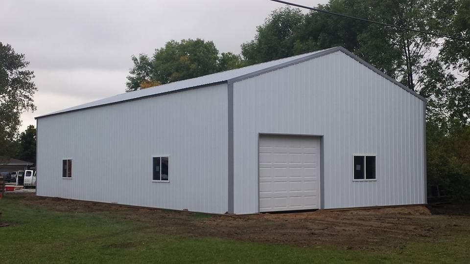 White metal building with a garage door and two windows, set in a grassy area with trees.
