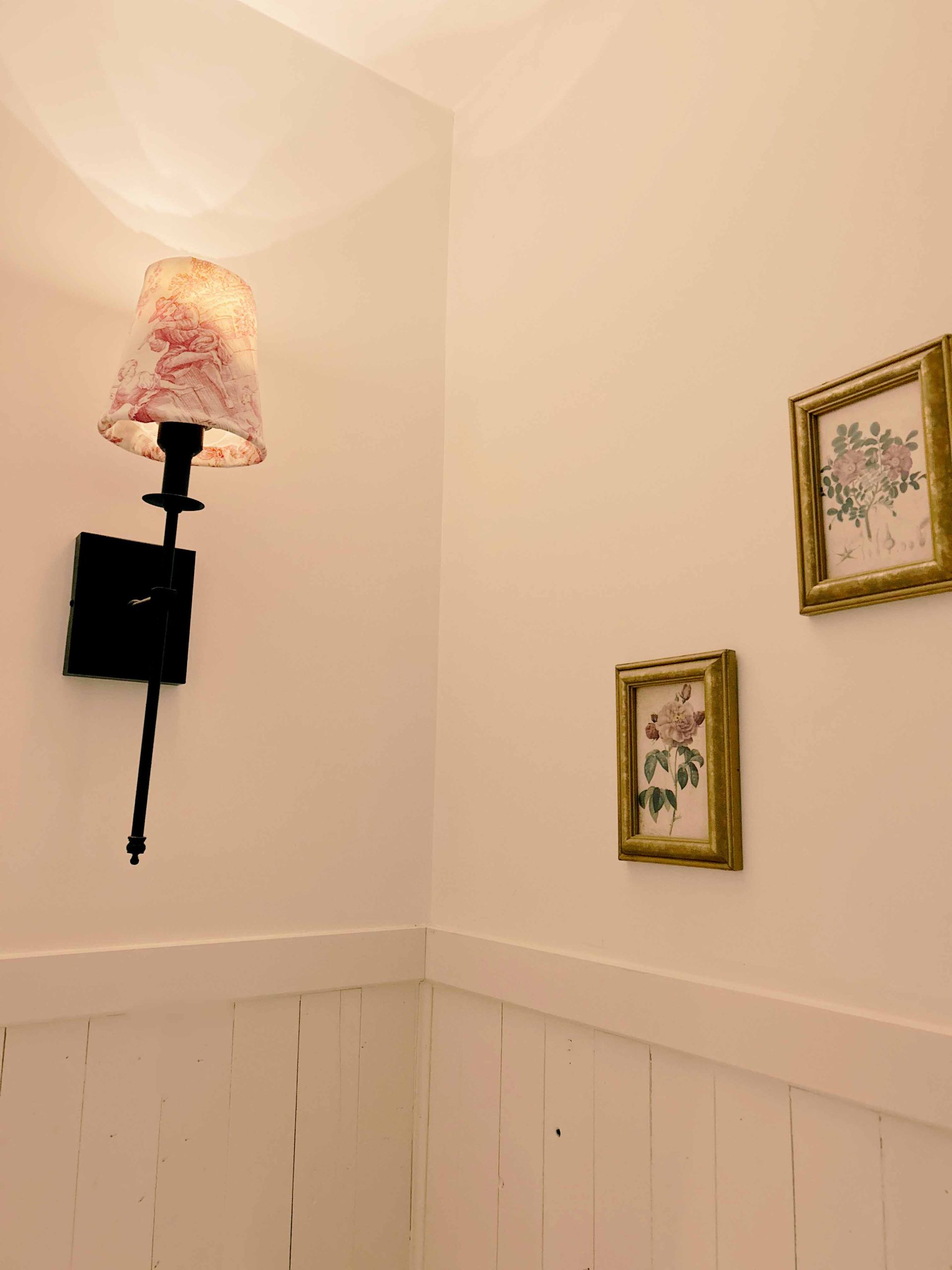 Applique murale noire avec abat-jour rose à motifs floraux sur un mur blanc orné de deux estampes florales encadrées.
