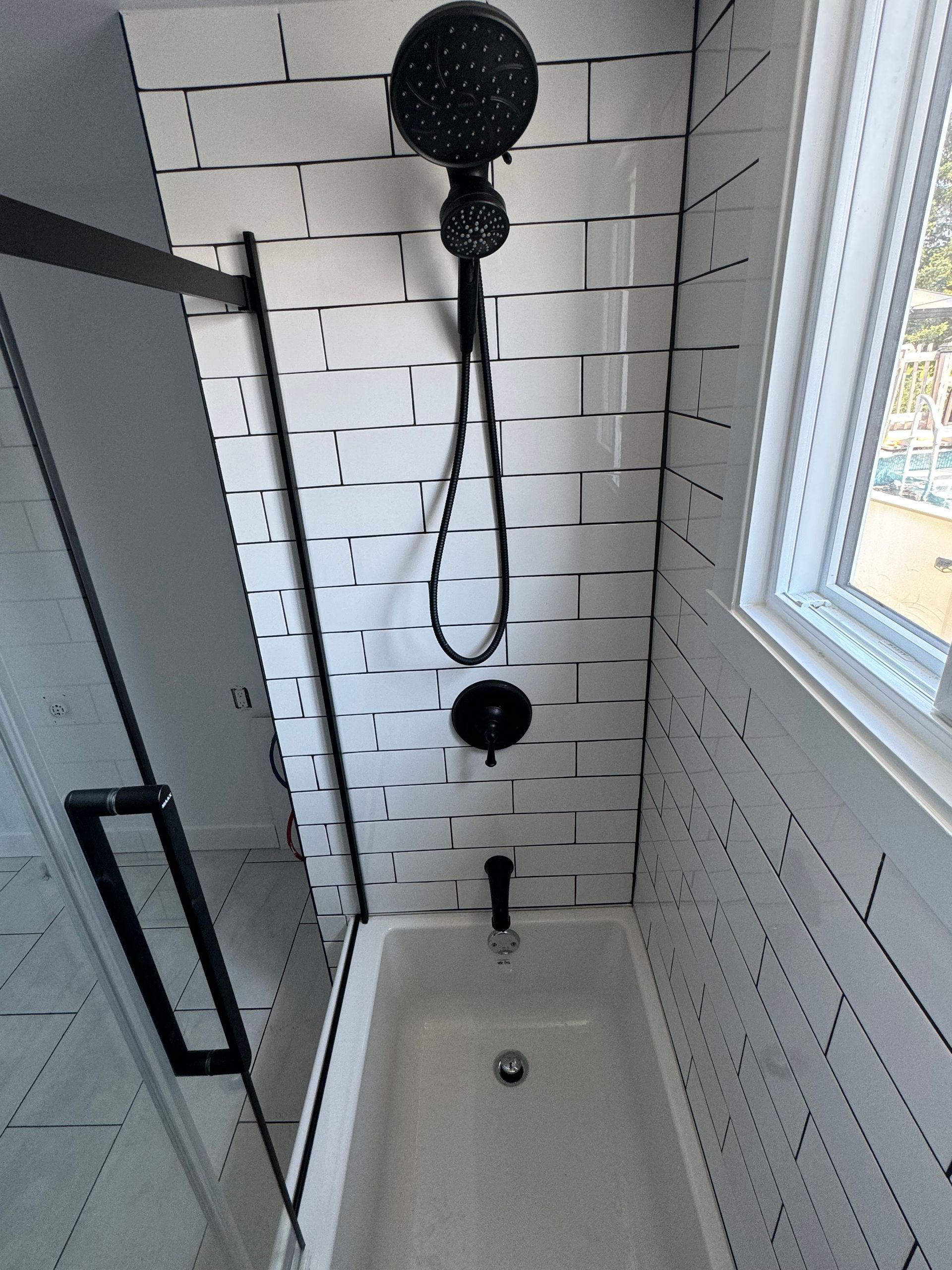 Petite salle de bains avec carrelage blanc imitation brique, robinetterie noire, porte en verre et petite fenêtre.
