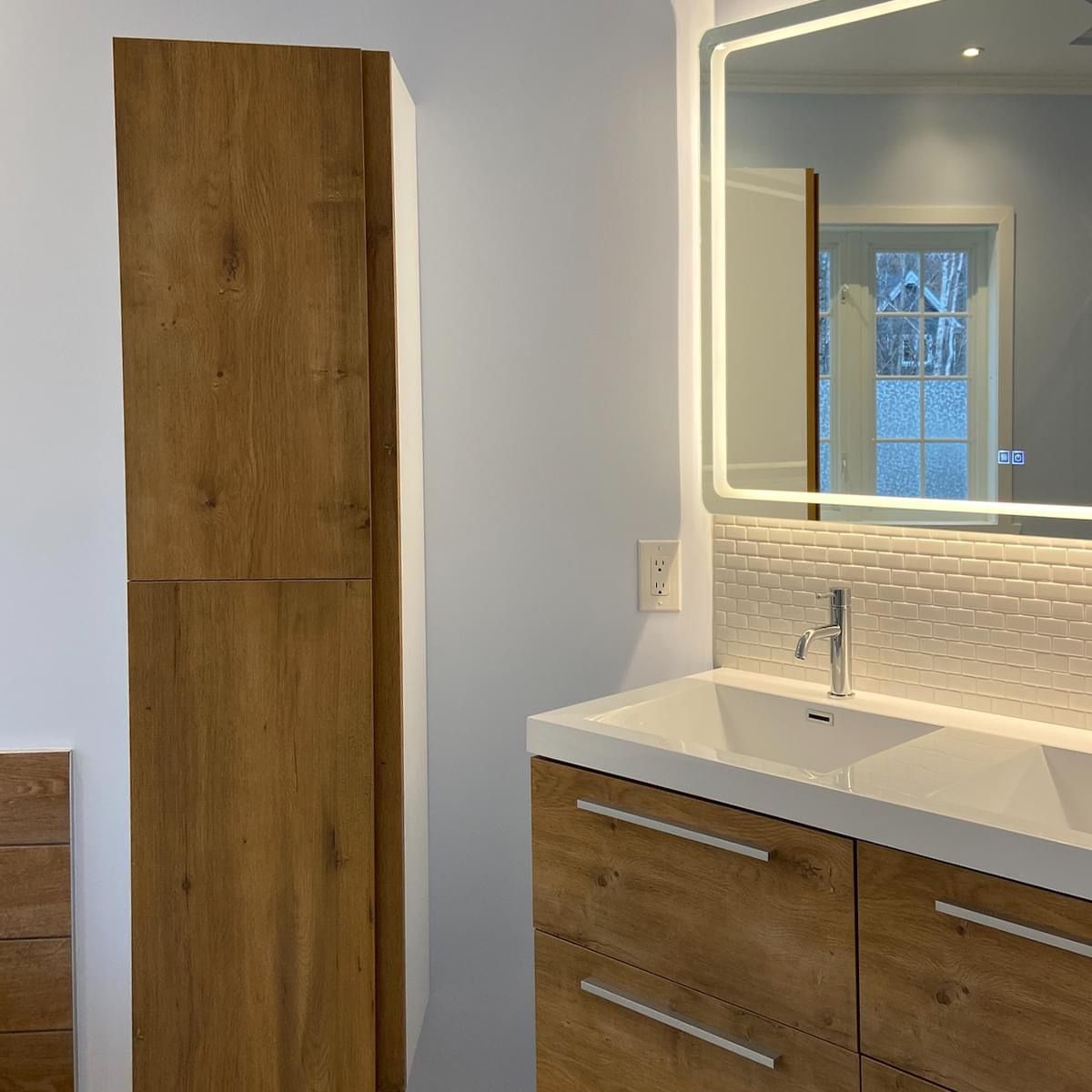 Salle de bains avec meubles en bois, comptoir blanc, miroir éclairé et crédence en mosaïque.