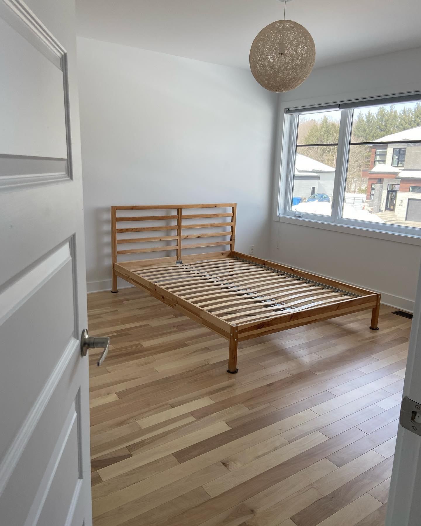 Chambre vide avec parquet et cadre de lit en bois, une fenêtre et un luminaire décoratif.