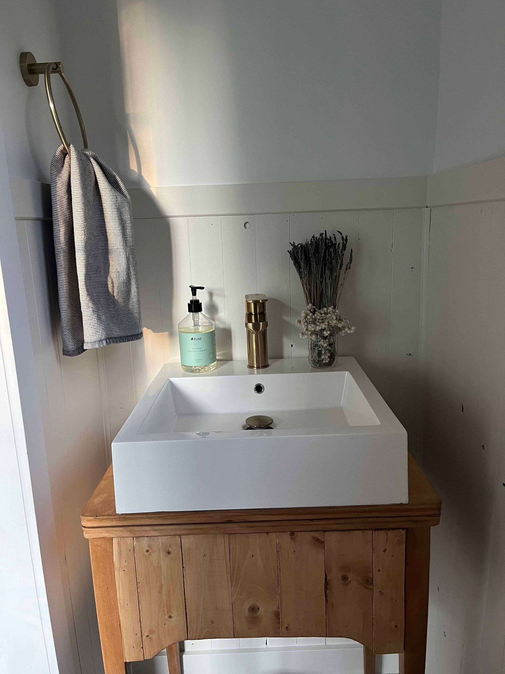 Lavabo rectangulaire blanc sur meuble en bois, robinet doré, savon, lavande, serviette rayée suspendue.