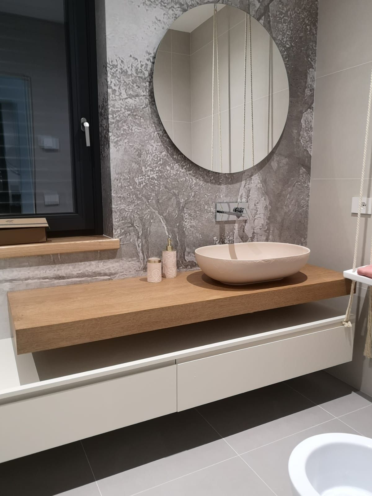 Mobile bagno moderno con piano in legno, specchio rotondo, lavabo e parete grigia con finitura testurizzata.
