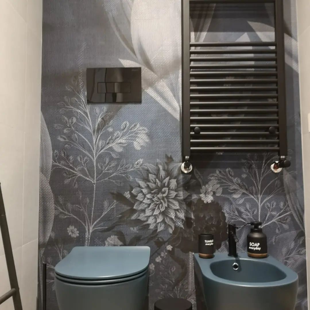 Bagno moderno con carta da parati a motivi floreali, WC e lavabo blu e tende scure alle finestre.