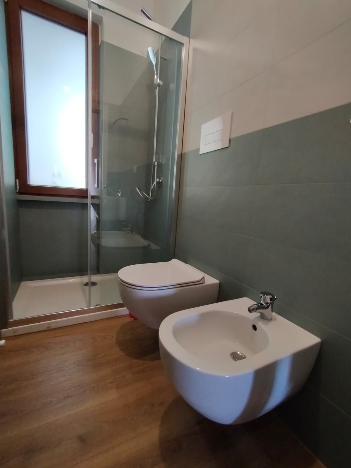 Bagno moderno con WC, lavabo, doccia e pavimento effetto legno vicino a una finestra satinata.