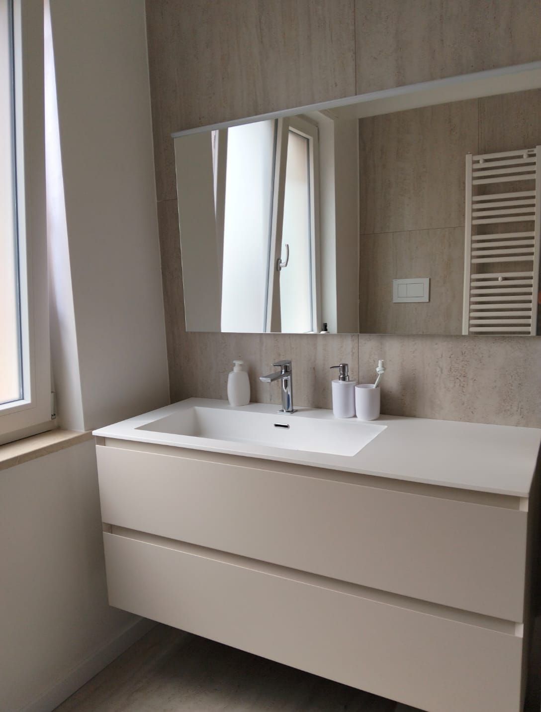 Mobile bagno moderno con lavabo bianco, ampio specchio e pareti rivestite con piastrelle beige vicino a una finestra.
