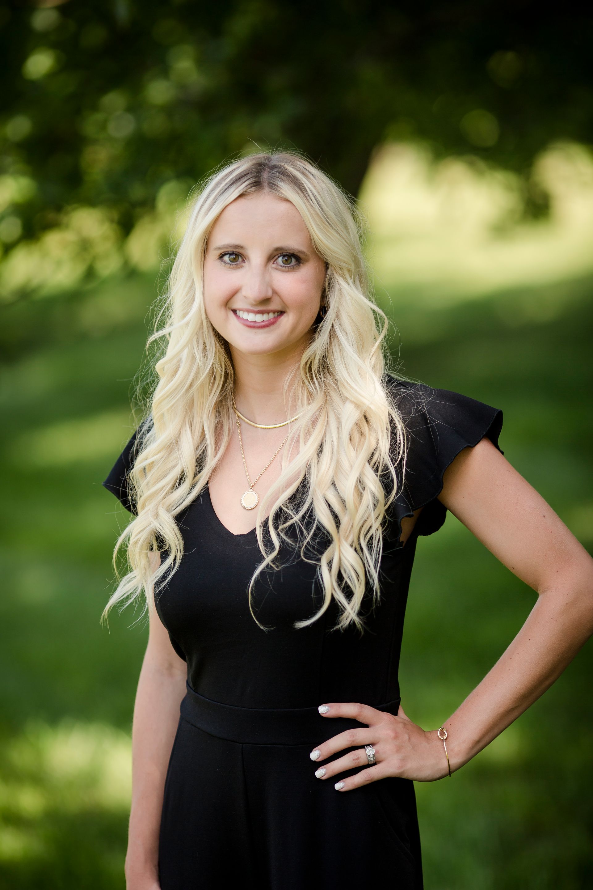 A smiling person with long blonde hair, wearing a black top, poses outdoors against a blurred green background.
