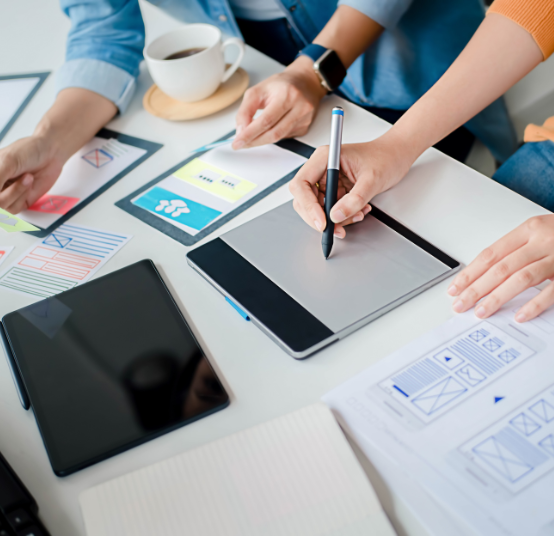 Designers collaborate at a desk, sketching website wireframes on paper, a tablet, and a digital drawing pad.