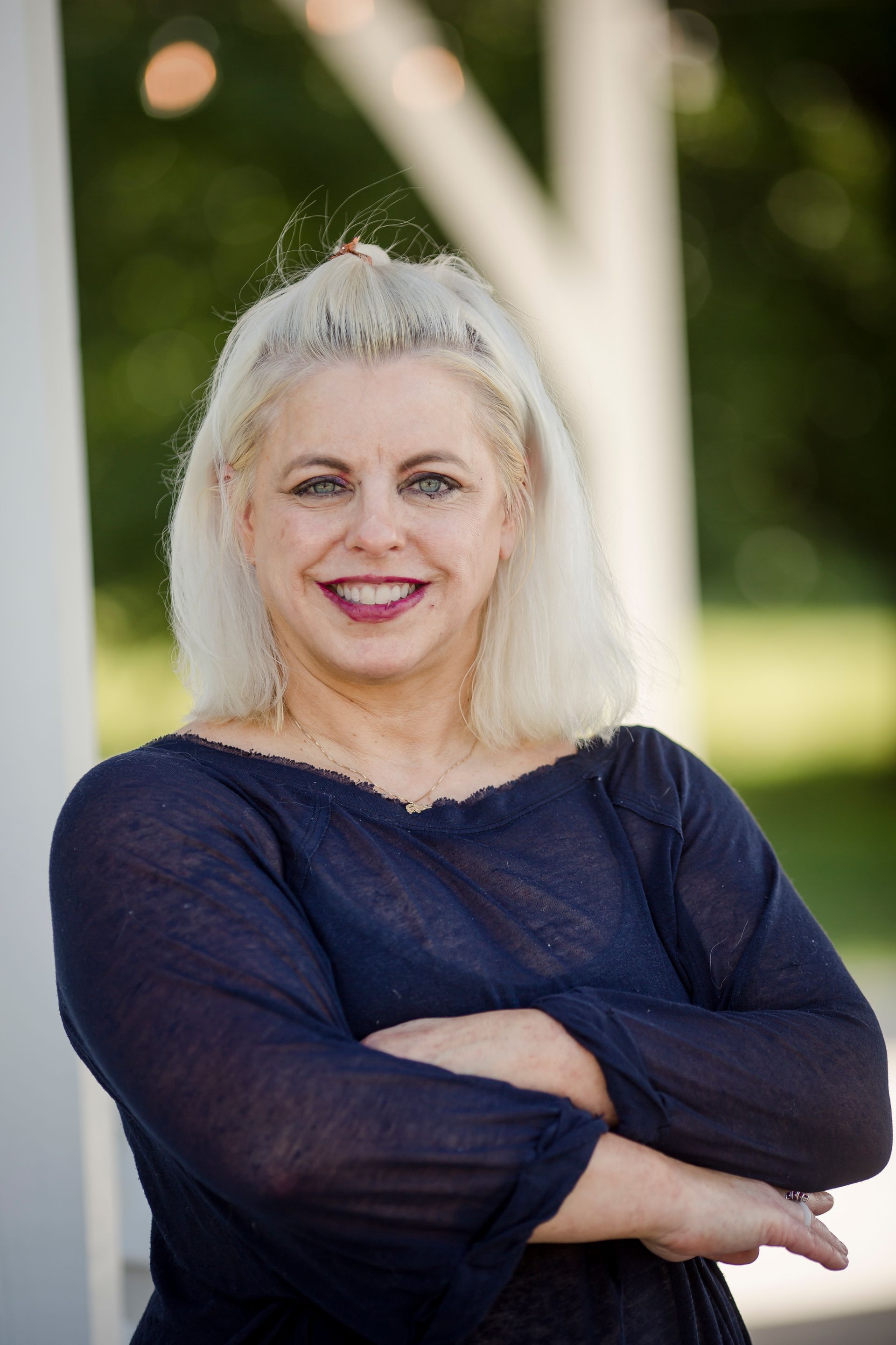 A woman with light hair wearing a navy blue top stands with her arms crossed outdoors.