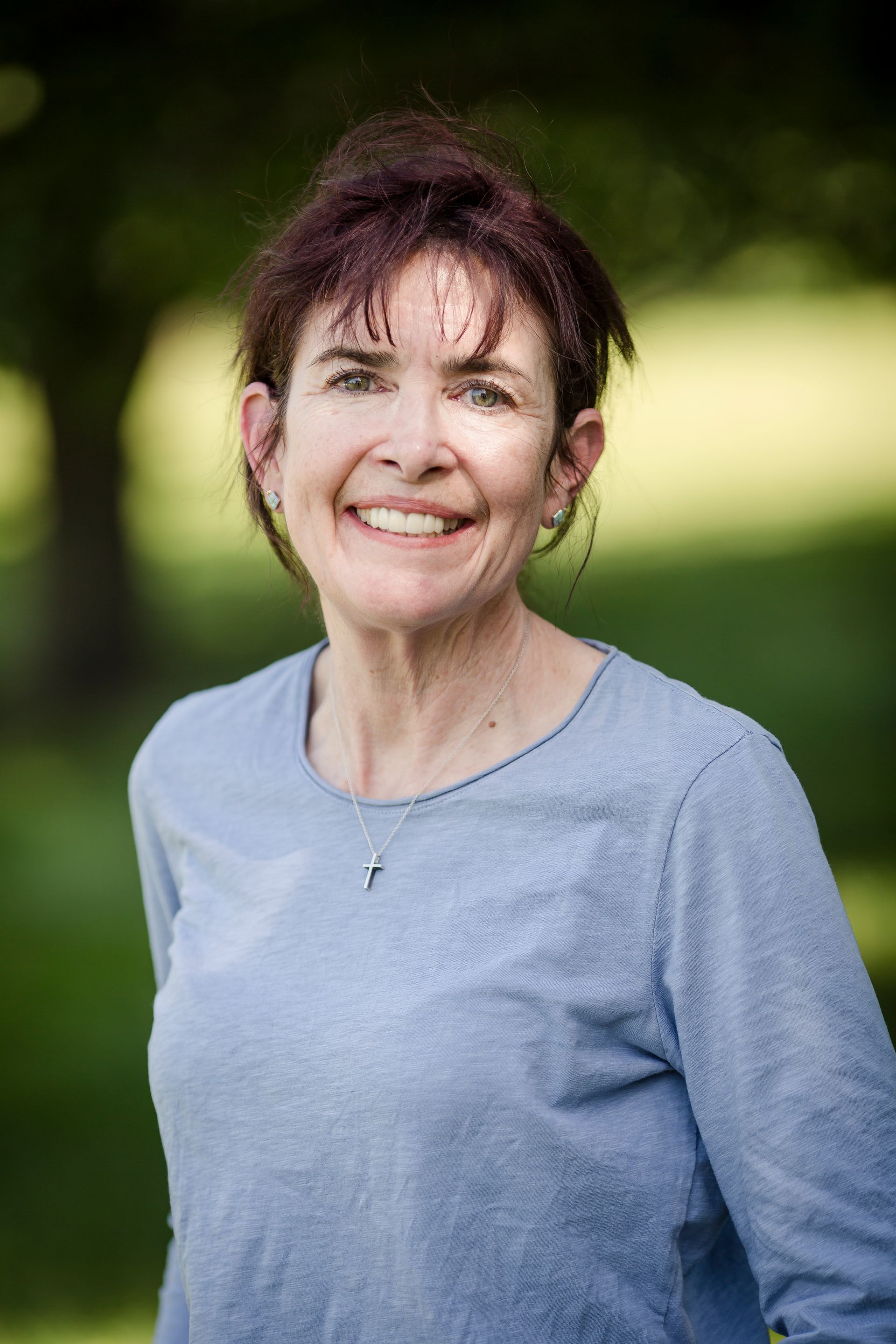 A person with shoulder-length reddish-brown hair smiles warmly against an out-of-focus green background.