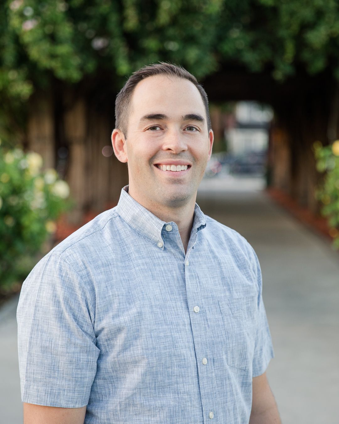 A smiling person in a blue button-down shirt standing outdoors in front of a blurred garden path.