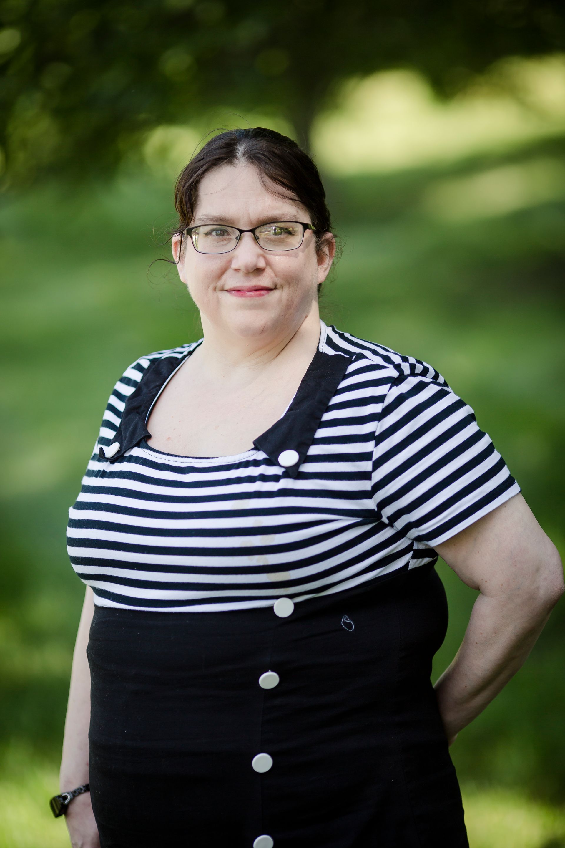 A person wearing glasses and a black-and-white striped top with a buttoned black skirt, posing outdoors in greenery.