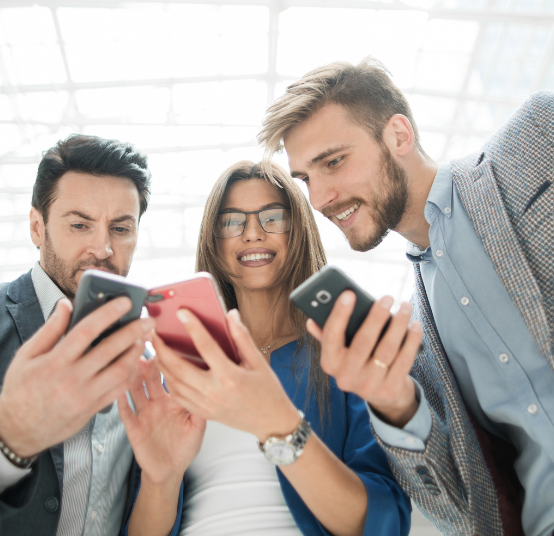 Three people in business attire stand closely together, looking down and smiling at their individual smartphones.