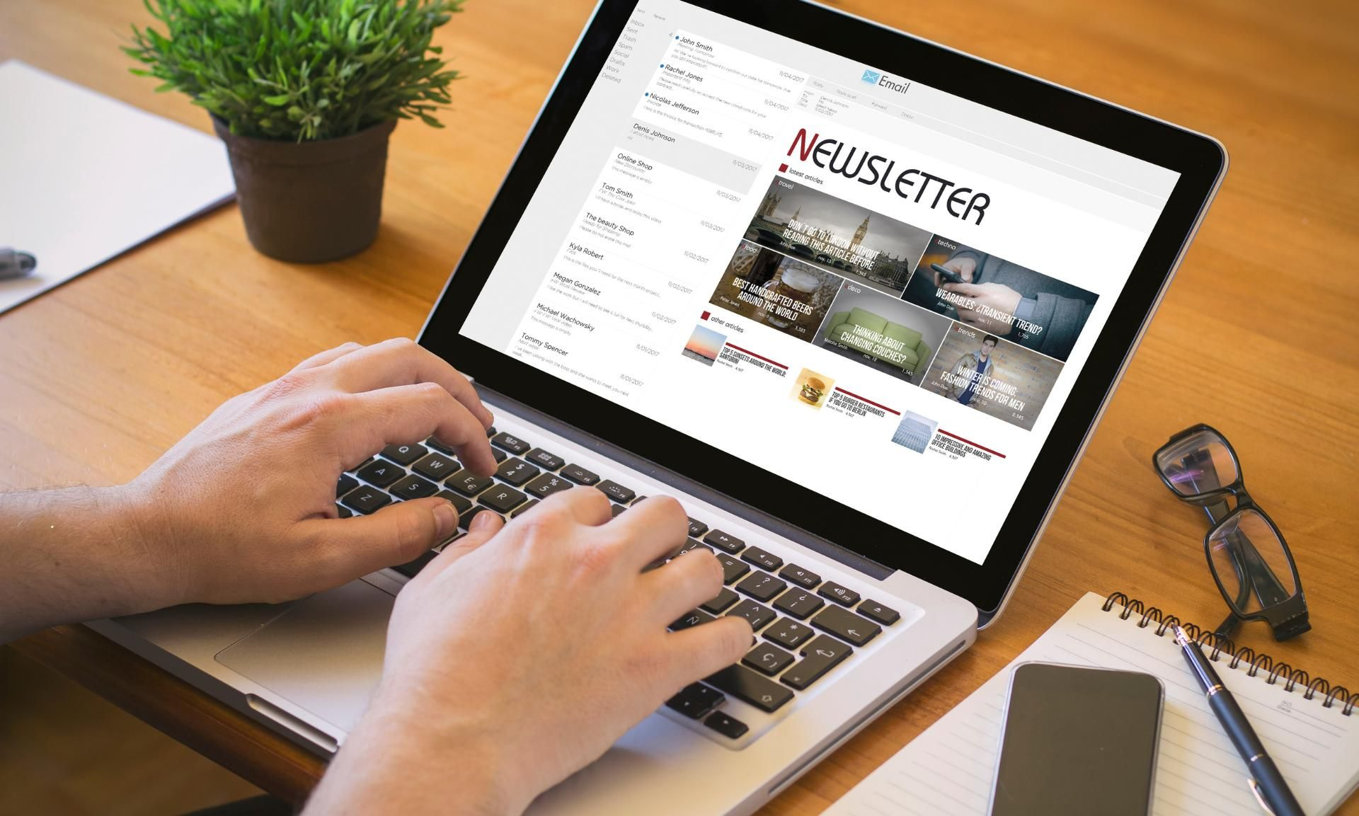 A person typing on a laptop displaying a newsletter page on a desk with a plant, notebook, and glasses.