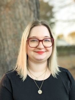 A person with shoulder-length blonde hair and red glasses smiles in front of a tree. They wear a black top and a necklace.