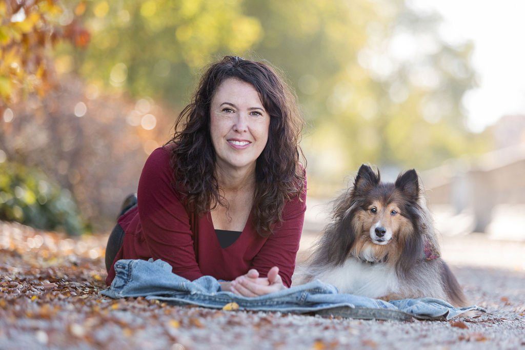 Woman with curly hair smiles, lies on a blanket next to a fluffy Sheltie dog in an outdoor setting with fall foliage.
