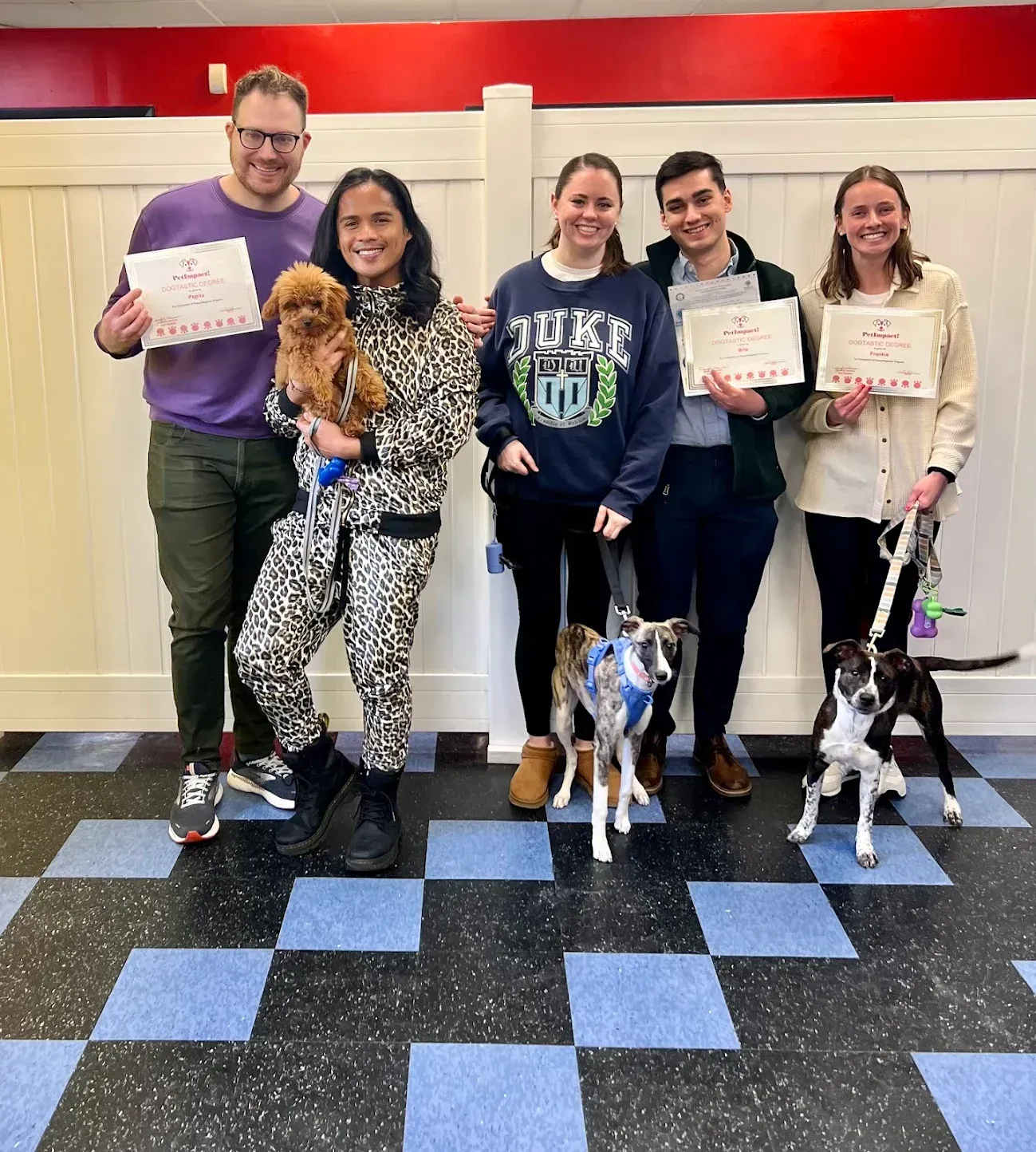 Group of people with dogs holding certificates in front of a white fence.