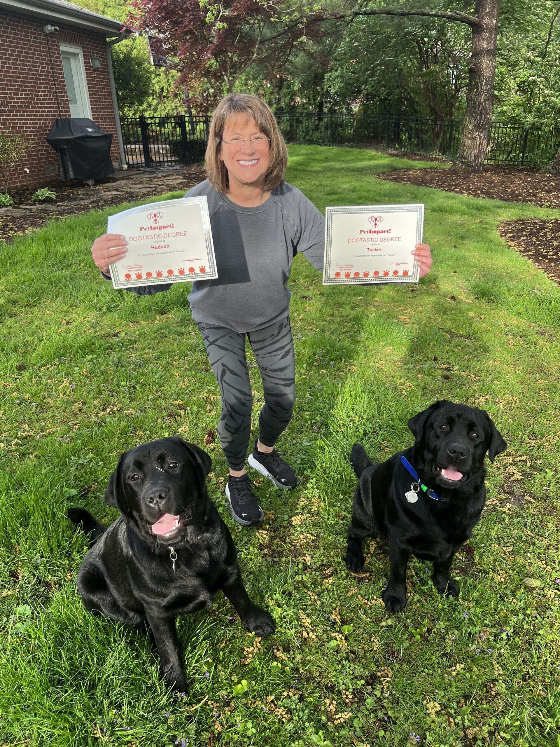 Woman in gray outfit holds two certificates, flanked by two black Labradors on a grassy lawn.