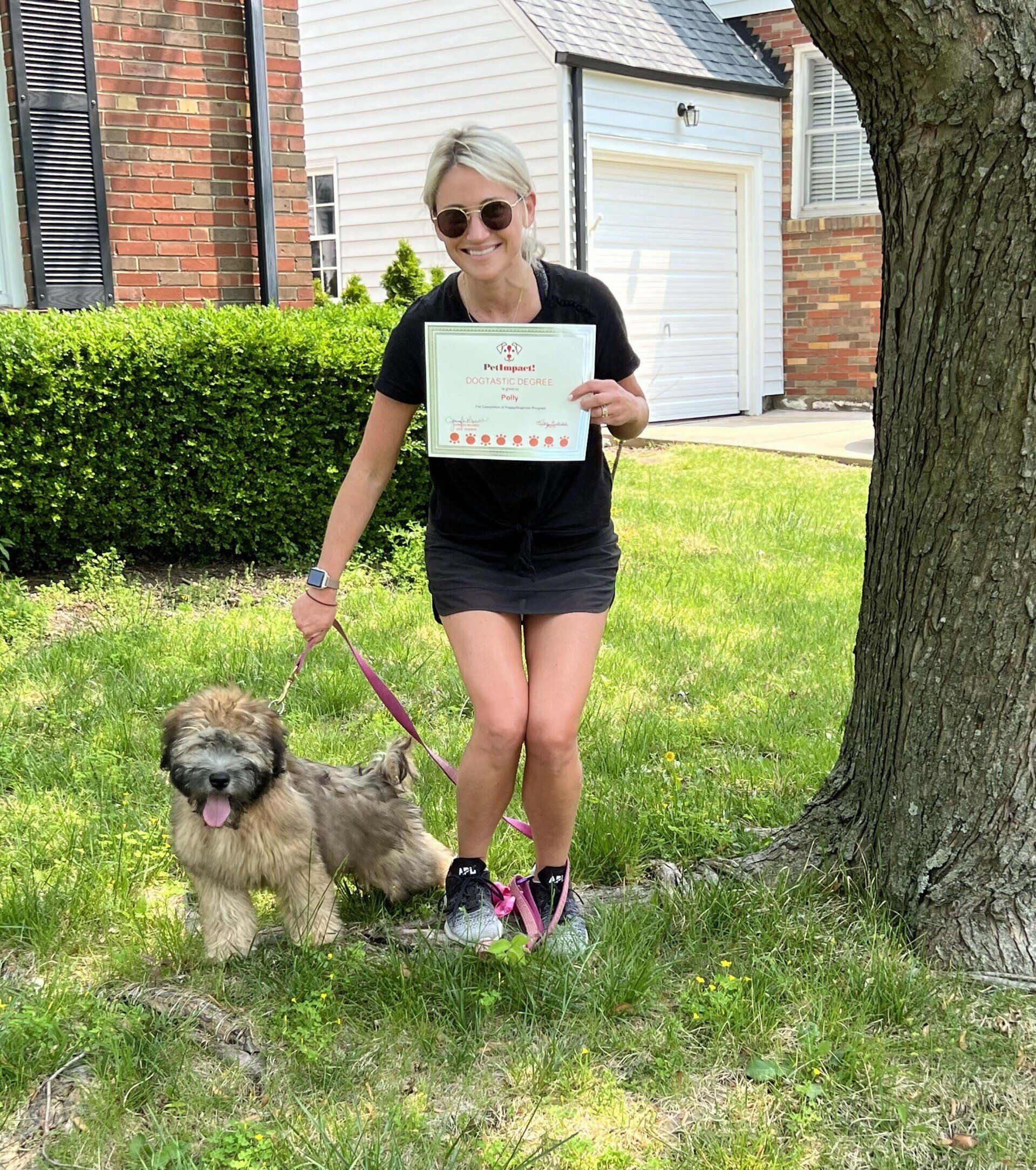 Woman holding a certificate, smiles at camera, and stands with a fluffy tan dog on a leash near a tree.
