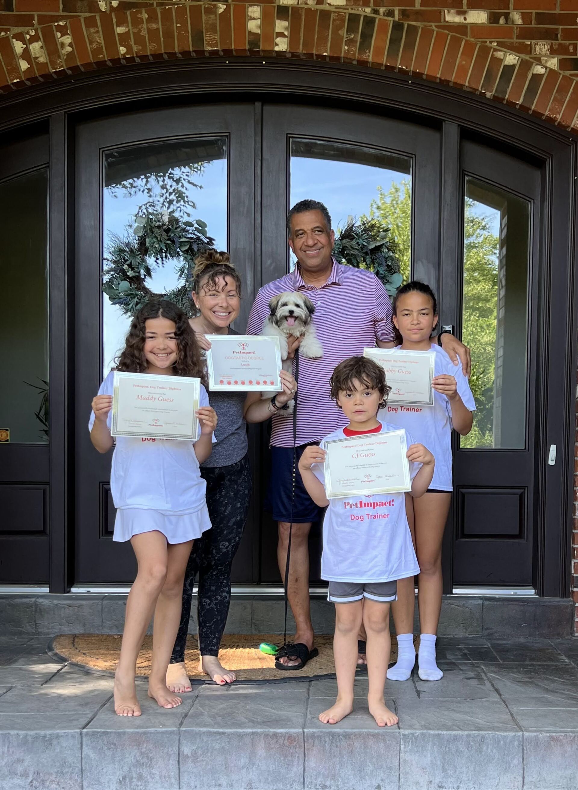 Family of five holding certificates, posing in front of a house with their small dog.