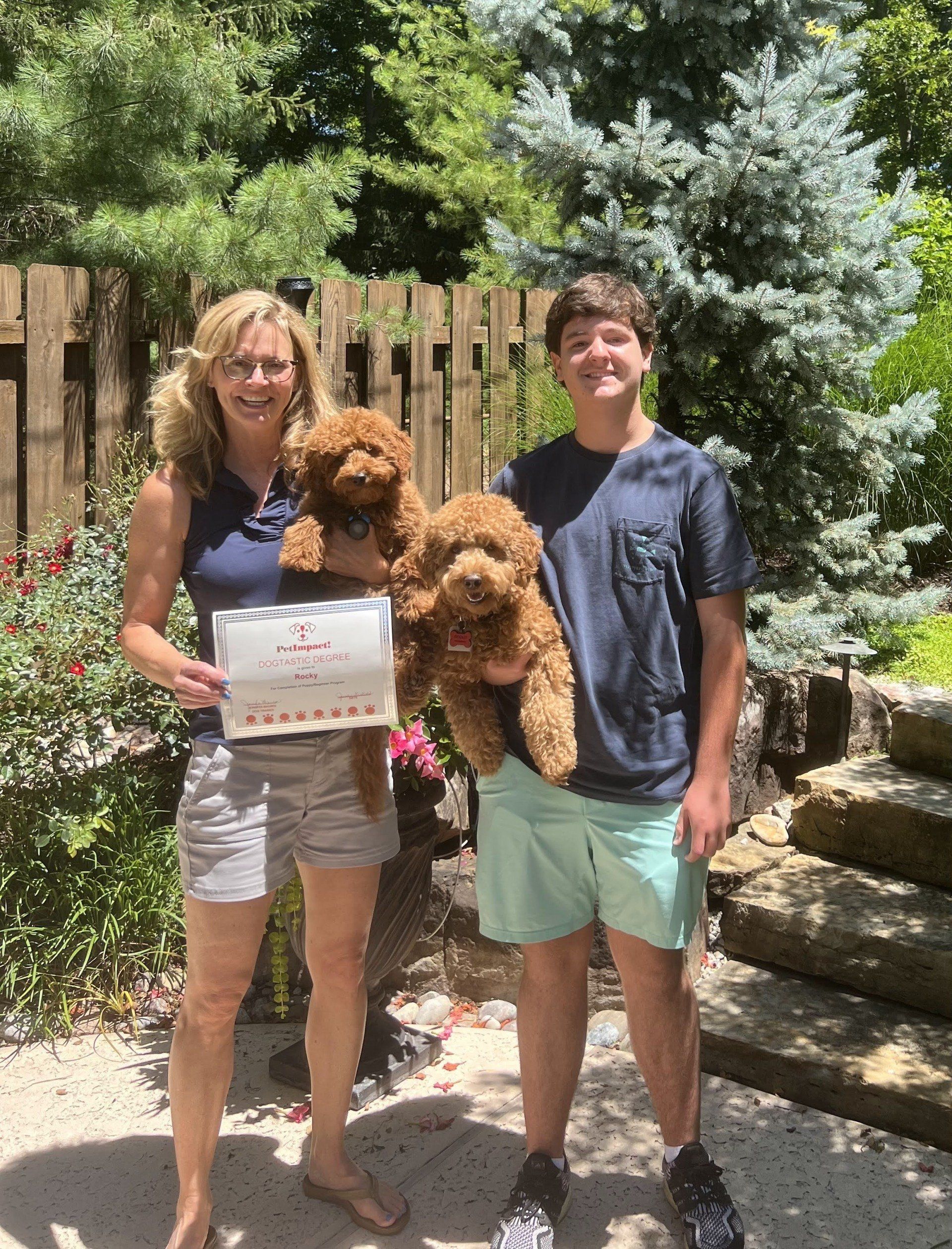 Woman and teen hold two brown dogs, standing outside, woman holding document.