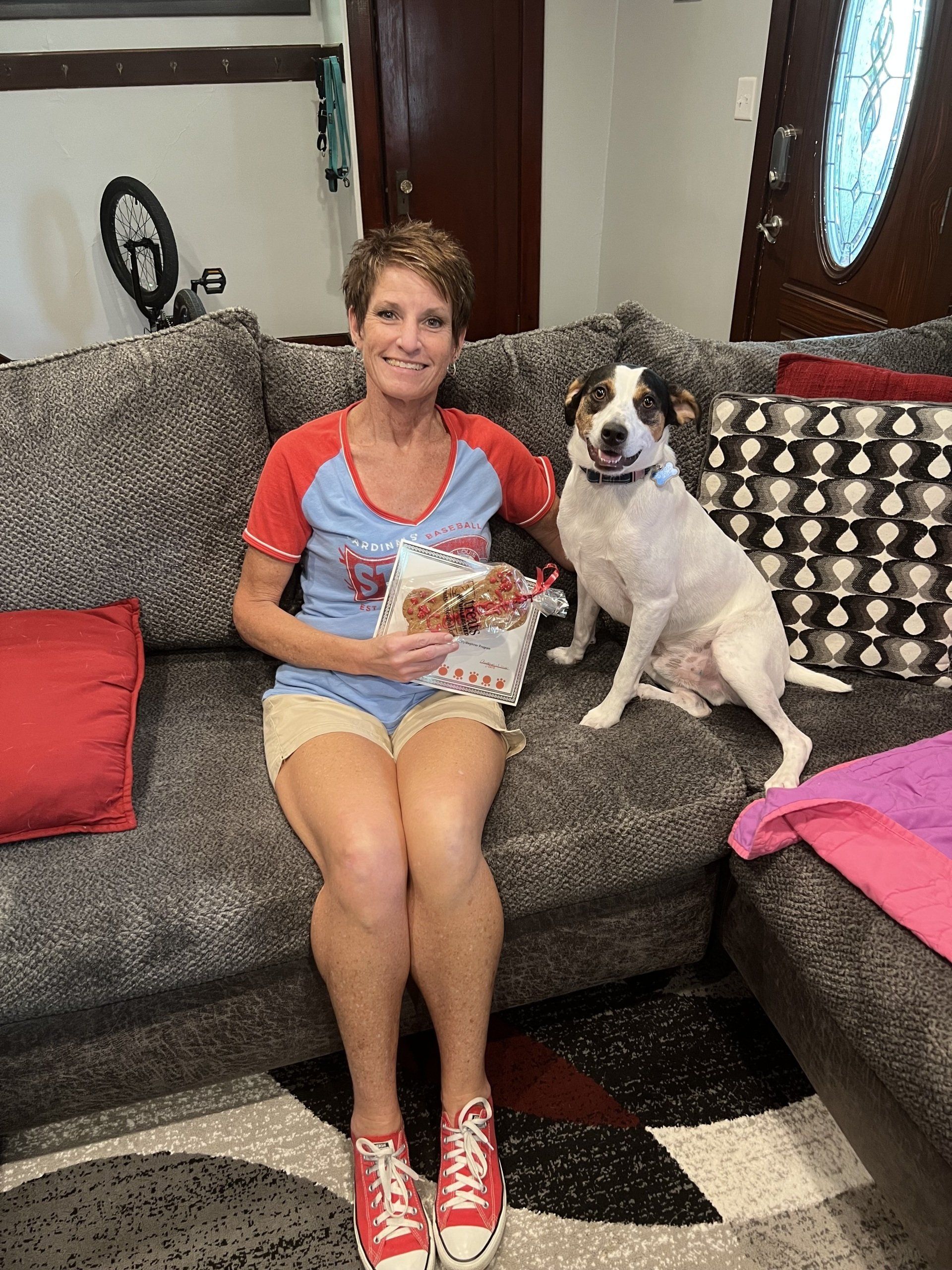 Woman and dog sitting on a gray couch. Woman holds a treat package, both smile.