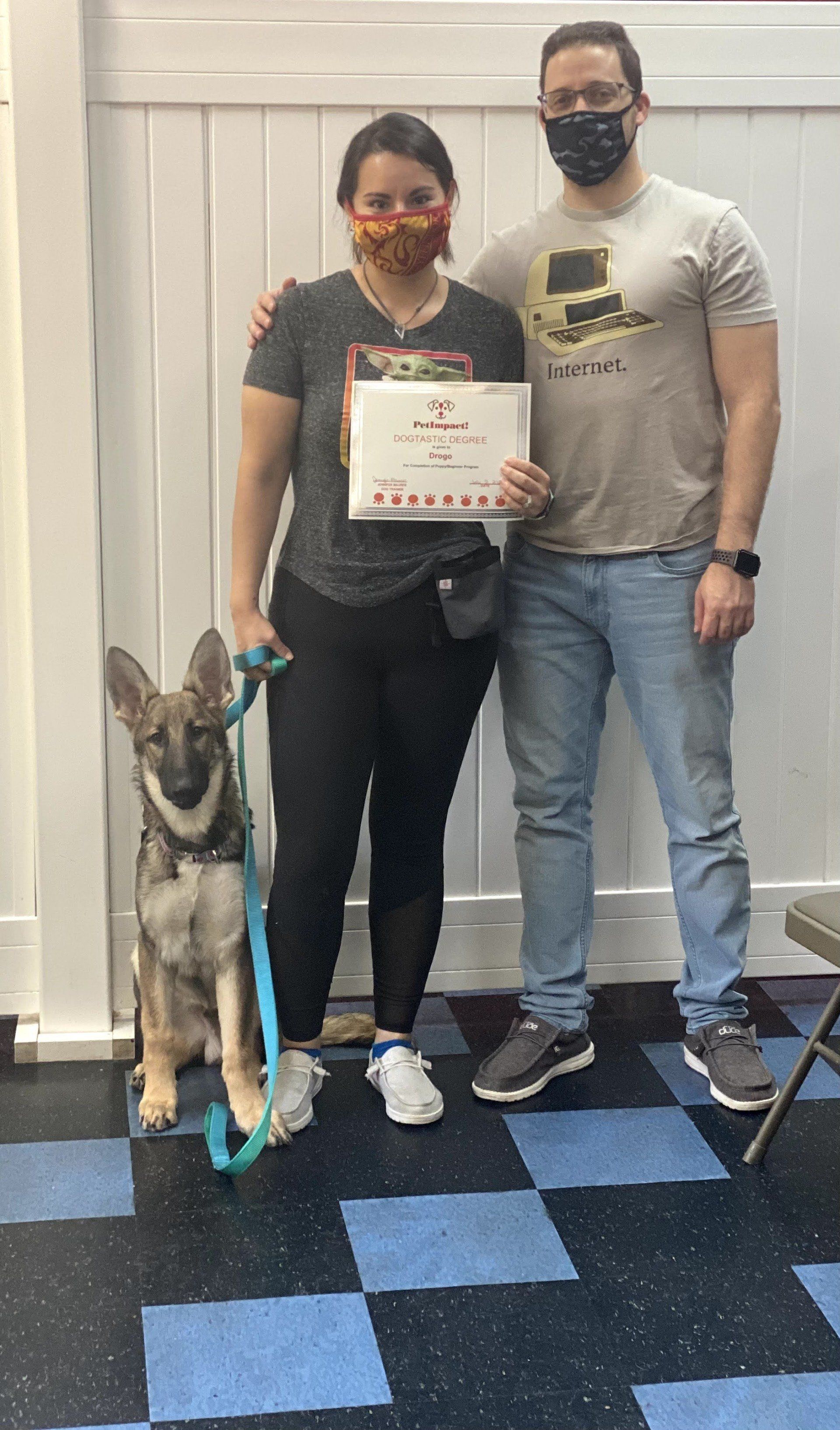 Woman and man with a dog holding certificate; inside, black, white, and blue checkered floor.