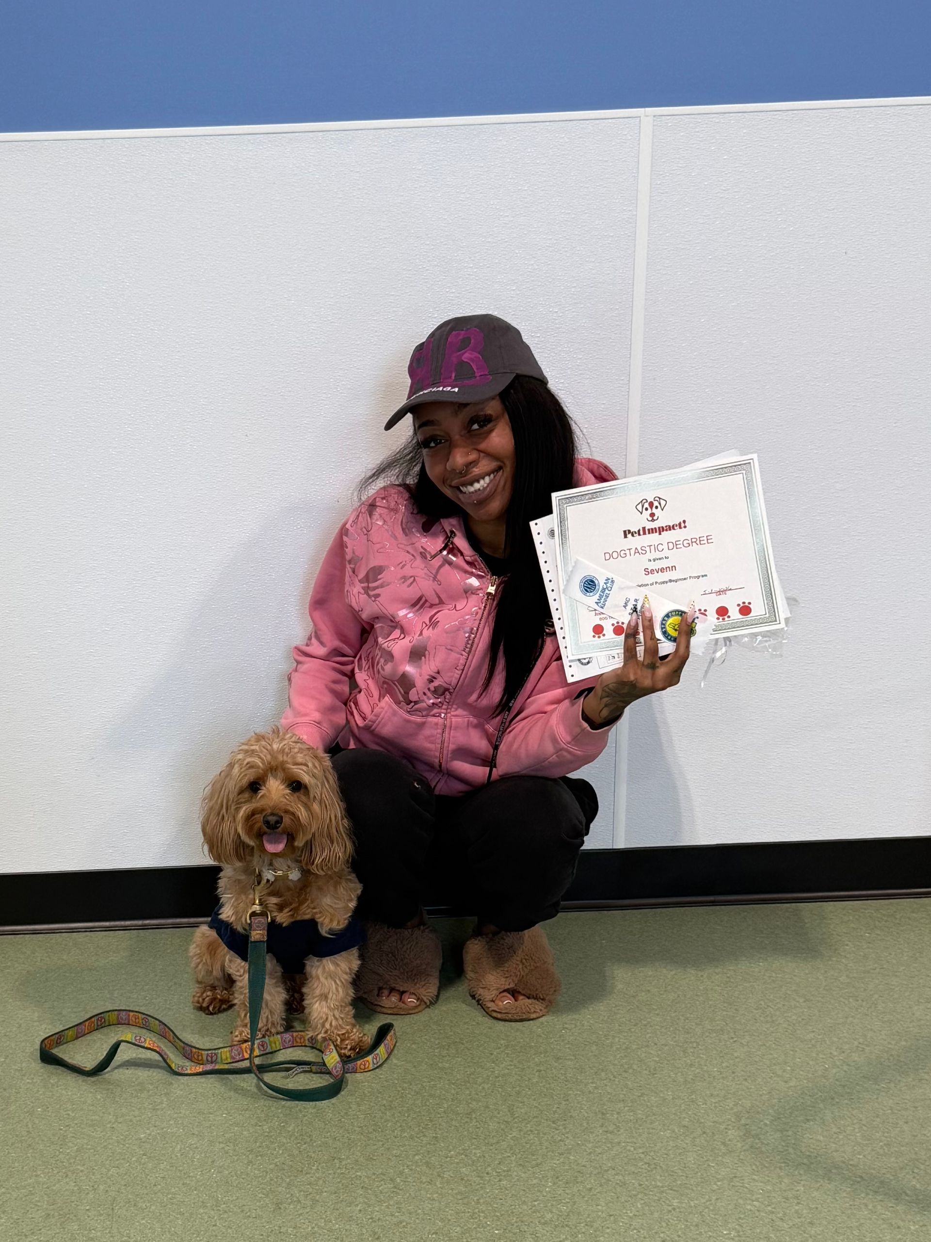 Person kneels beside a small dog, holding up a certificate against a white wall.