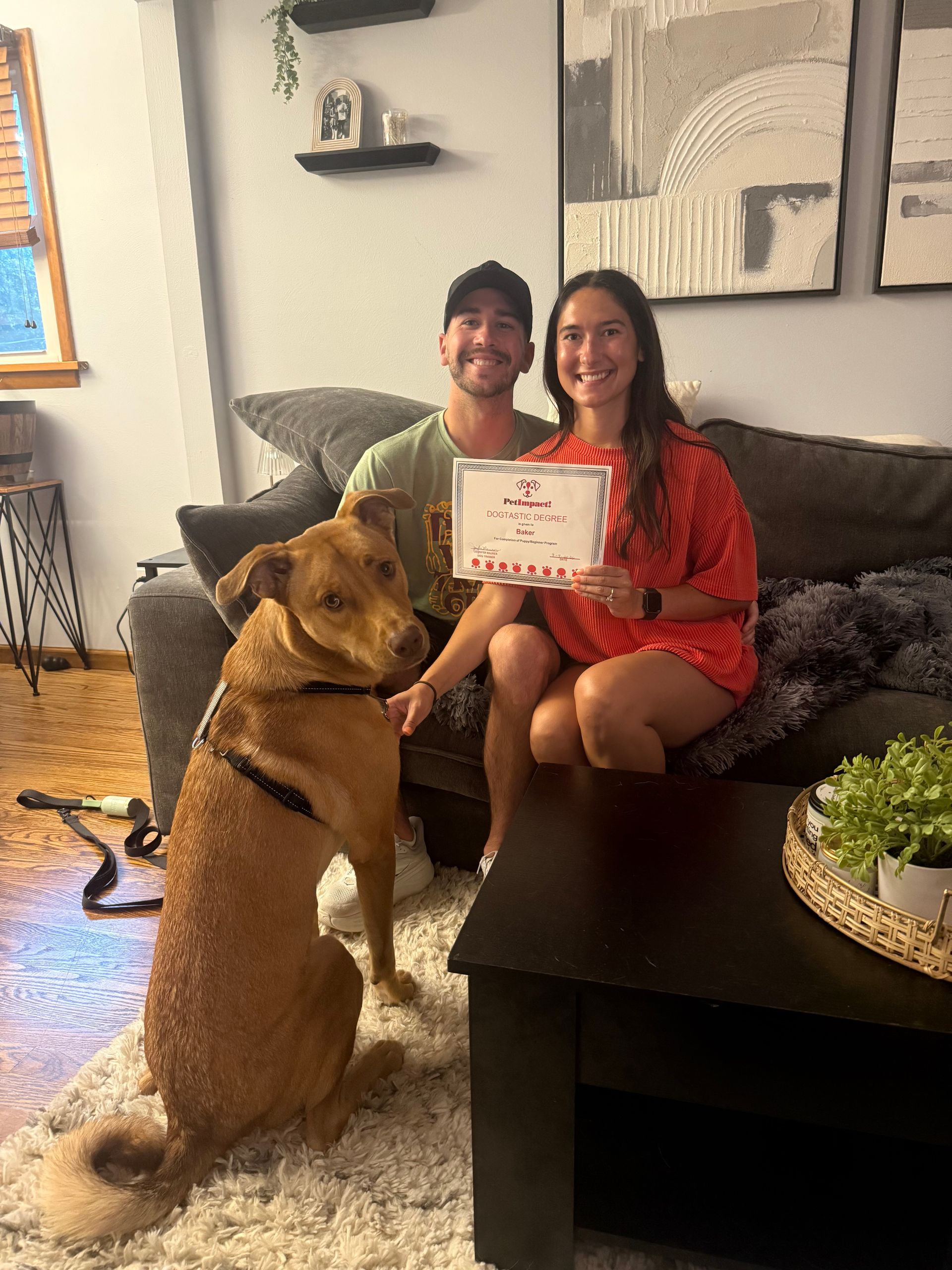 Man and woman on a couch holding a sign, with a large dog sitting in front of them in a living room.